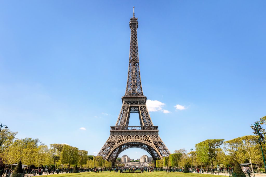 Eiffel Tower on a clear blue day with green trees and tourists in the foreground.