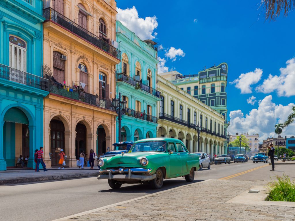 Classic green vintage car driving past colorful colonial buildings under a bright blue sky in Havana, Cuba.