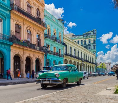 Image for Classic green vintage car driving past colorful colonial buildings under a bright blue sky in Havana, Cuba.
