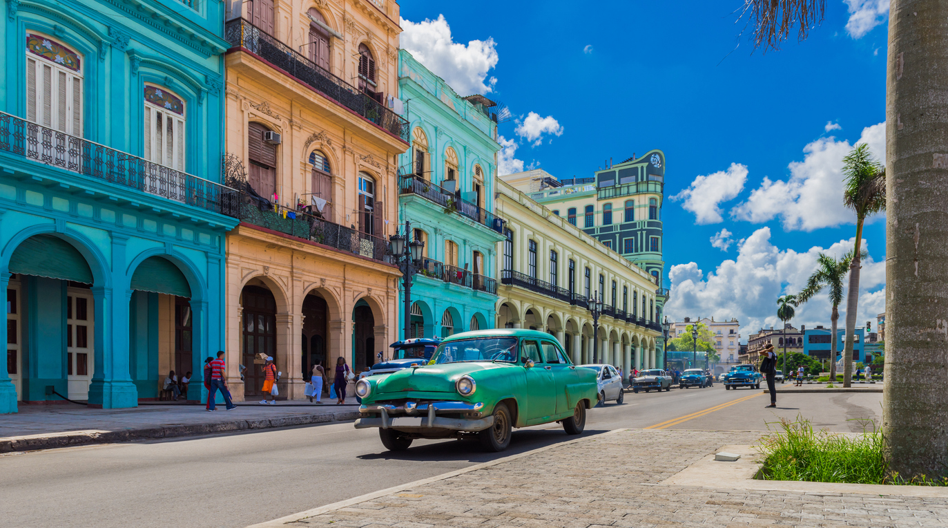 Image for Classic green vintage car driving past colorful colonial buildings under a bright blue sky in Havana, Cuba.